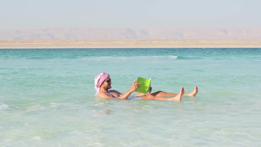 A male tourist effortlessly floats in the hypersaline turquoise waters of the Dead Sea while reading Jordan travel brochure book. He wears a red and white traditional Jordanian keffiyeh headscarf.