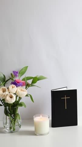 Minimal Christian still life featuring a Bible with a cross, burning candle and fresh flowers in a vase on a clean white background. Symbol of faith, prayer, hope, worship and spirituality