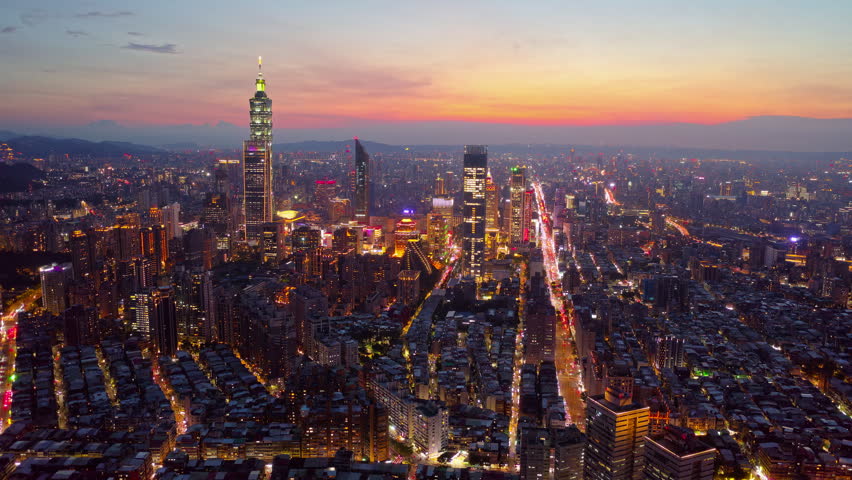 Aerial hyperlapse footage from dusk till night above Downtown Taipei, an international metropolis and the vibrant capital city of Taiwan, with 101 Tower standing out amid skyscrapers in XinYi Commercial District and city lights dazzling under dramatic twilight sky.