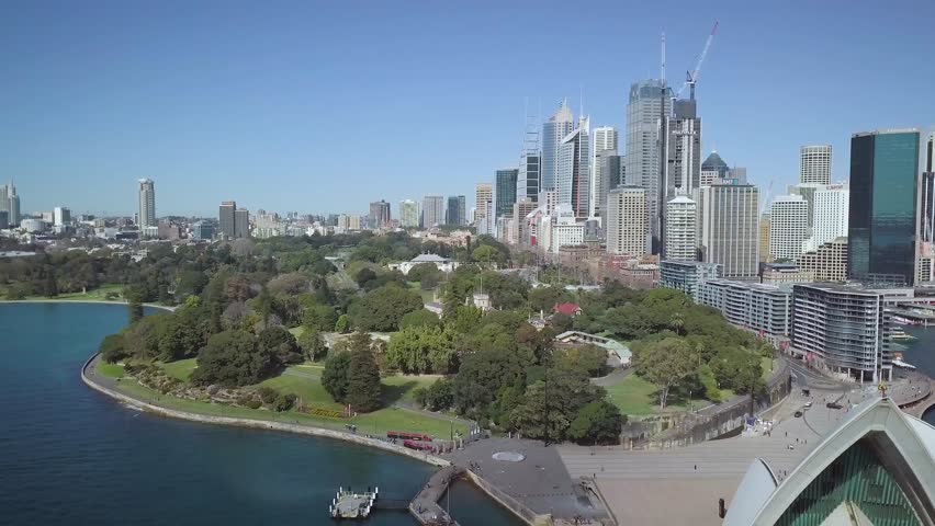 The Sydney Opera House sits in the Sydney Harbour near the Sydney Harbour Bridge on the coast of Eastern Australia. Drone footage captures its proximity to downtown Sydney.