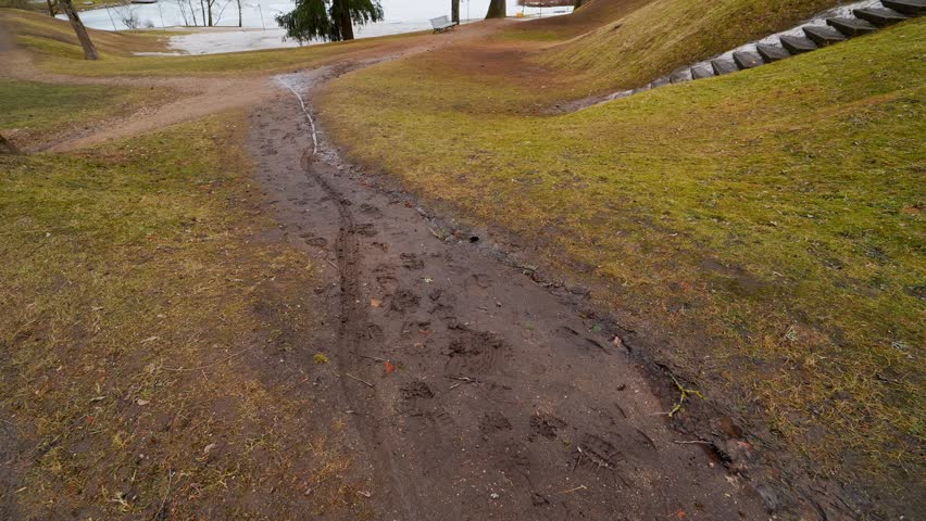 Muddy footpath in a quiet park during spring thaw, with melting snow, bare trees, and a partly frozen pond in the background.