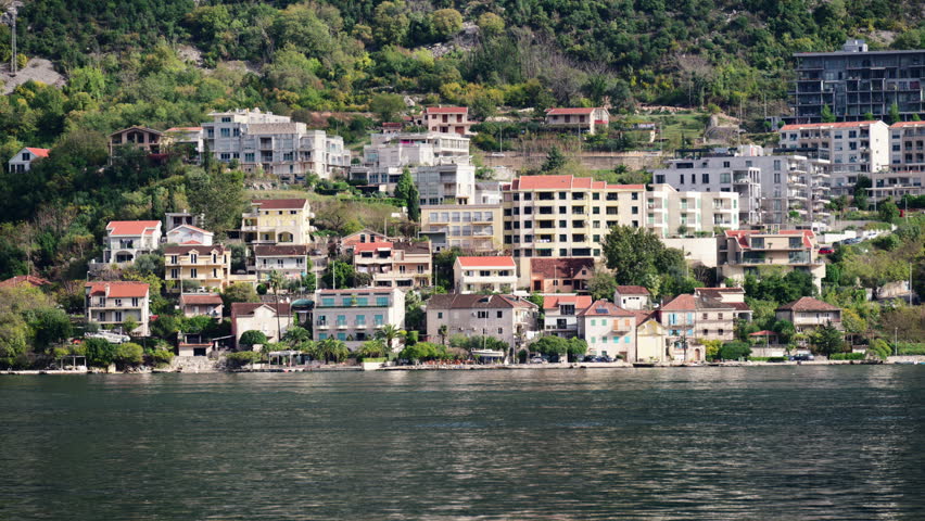 Houses and apartment buildings line the Bay of Kotor and green hillside. Boat passing by