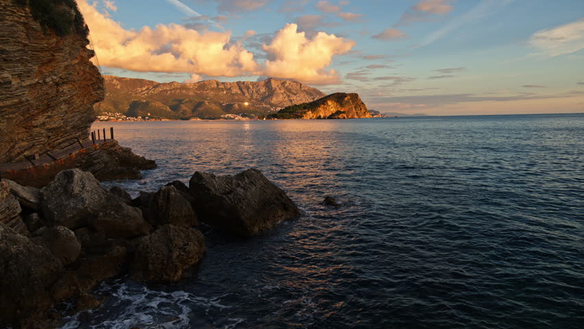 Adriatic Sea reflecting golden light from the setting sun over mountains and rocky shore. Budva, Montenegro
