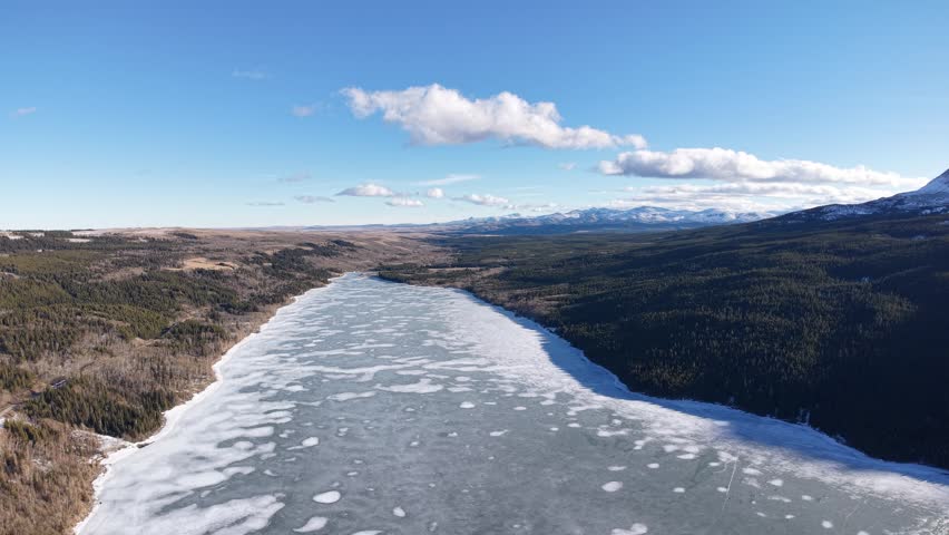 Wide aerial shot looking down on a long frozen river channel with melting ice floes stretching through a forested valley under a blue sky near Glacier National Park in Montana.