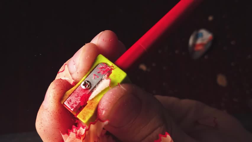 Close up of a hand sharpening a red pencil with a yellow sharpener, creating wood shavings.