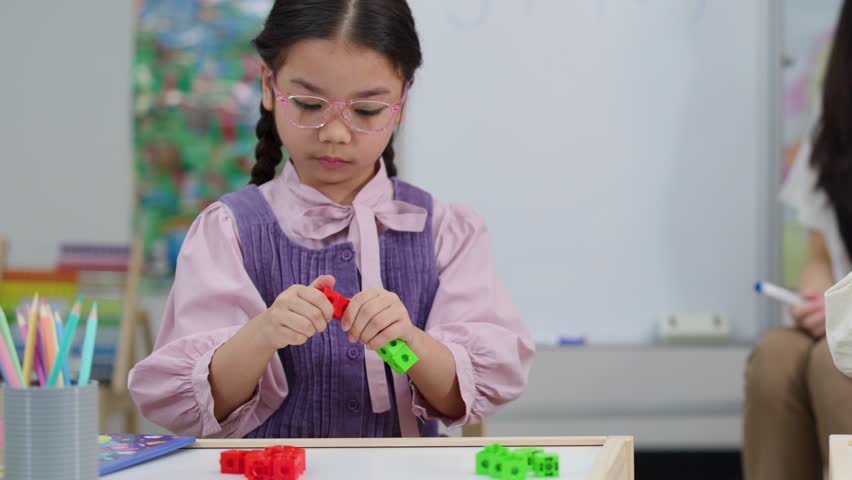 An elementary student wears glasses while connecting plastic blocks at a desk in a classroom