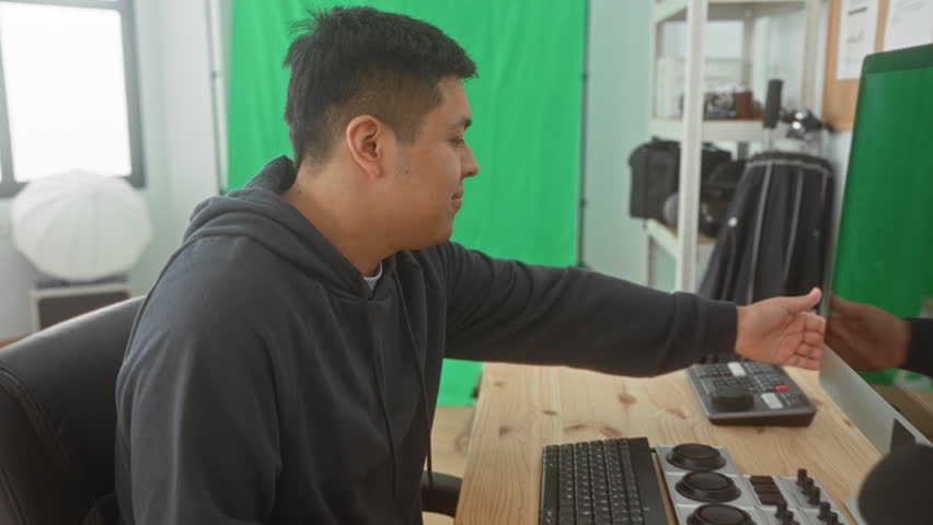 Man tapping smartphone screen while seated at desk with keyboard, microphone and green screen in studio; creative concentration.