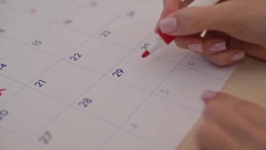 A hand writes the word Rent in red on a calendar date while holding a marker. The action suggests organization and planning related to rental payments.