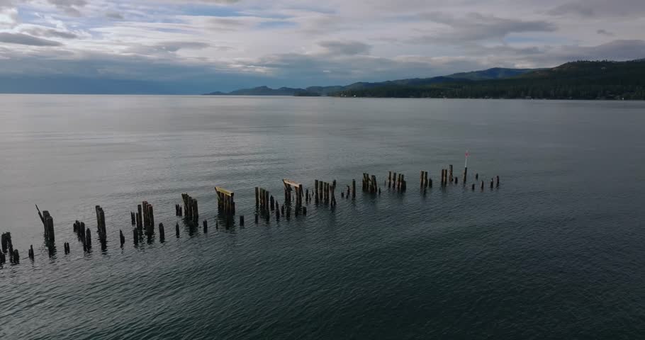 Row of wooden posts emerging from calm lake water, repeating pattern across the reflective surface under cloudy sky, aerial drone landscape, Flathead Lake Montana USA