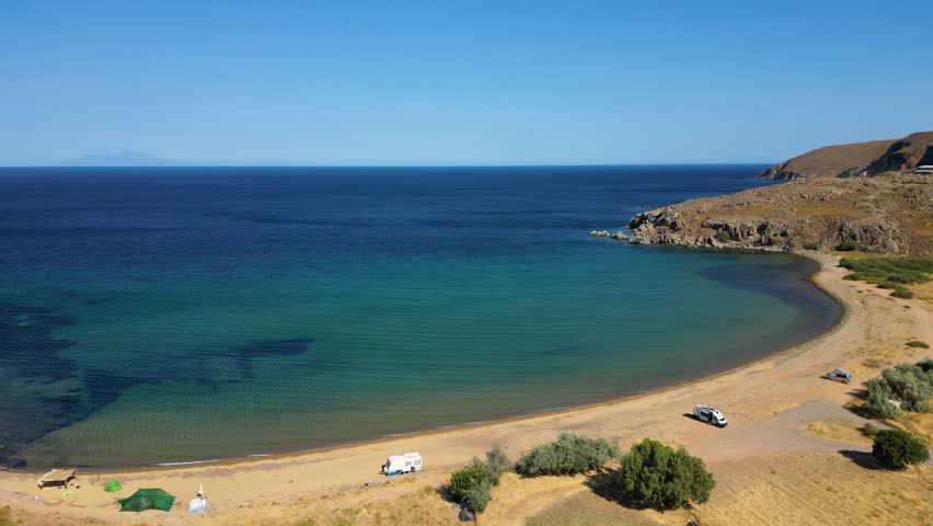 Aerial view of the secluded Karvounolakas beach on Limnos Island, Greece, featuring golden sands, turquoise waters, and camper vans parked for wild camping along the rugged Aegean coastline.