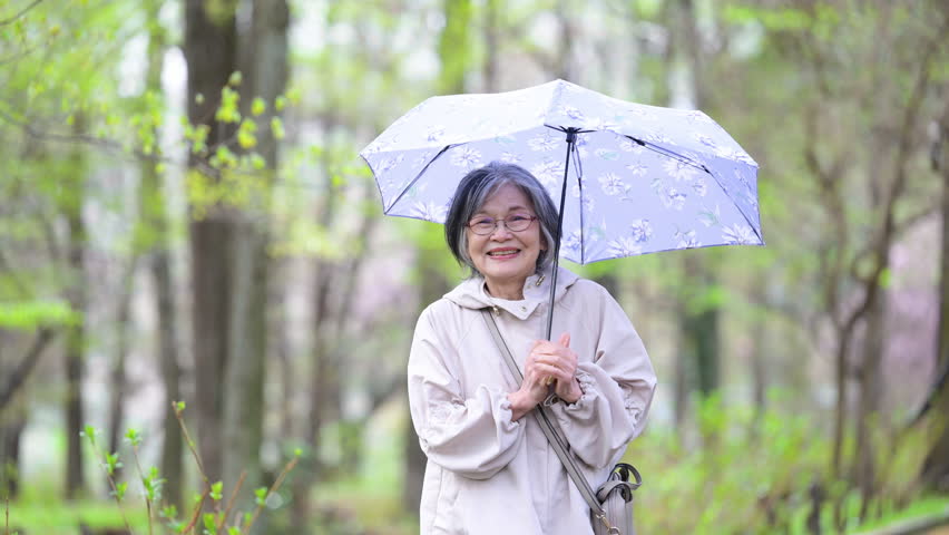 Video of an elderly woman taking a leisurely stroll with an umbrella, portraying a calm and healthy active senior, imagery of the rainy season, rain and parasols	