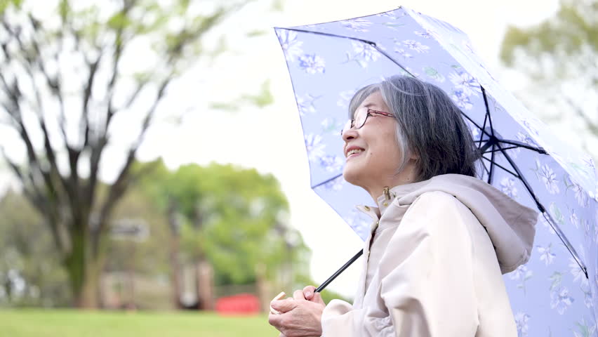 A close-up video of an active senior woman, looking up calmly and healthily while enjoying a walk in the rain or during the rainy season, holding an umbrella	