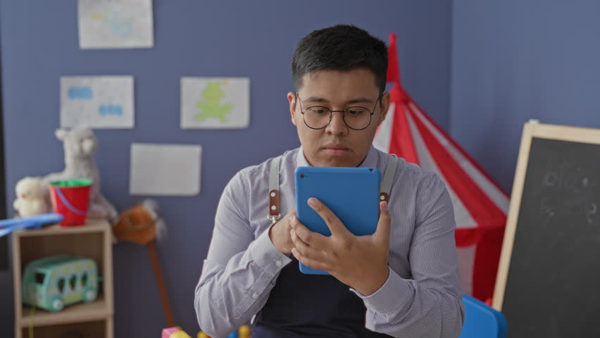 Man teacher holds blue tablet and taps screen in school building amid toys and play tent, wearing apron and glasses; concentration learning.