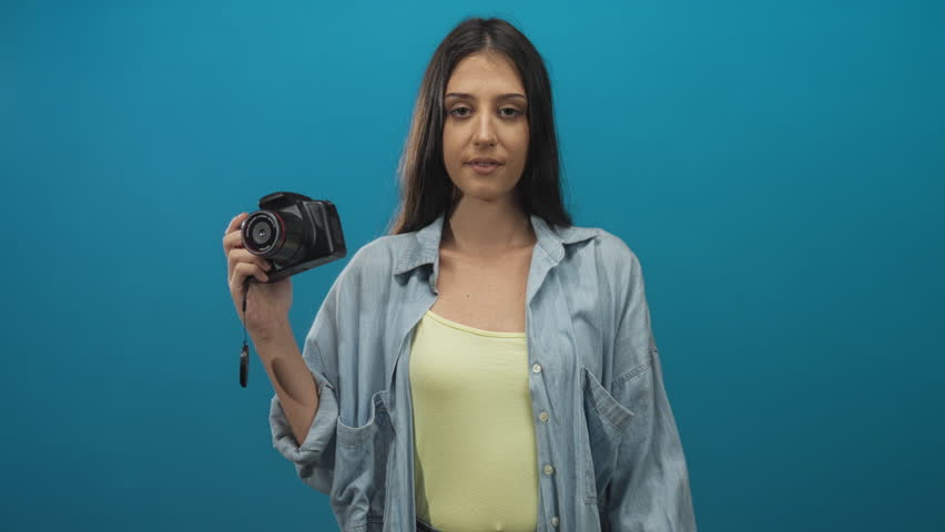 Young woman holding dslr camera with hand on chest and open mouth, wearing yellow tank top in teal studio; surprise discovery.