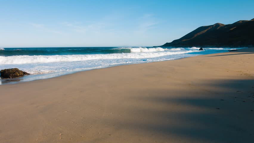 Waves crash along sandy shoreline at Point Mugu State Park with coastal mountain rising in background under clear blue sky creating scenic California beach landscape