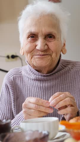 Vertical video of happy smiling senior woman with bright open smile and sparkling eyes eating at table and drinking tea. Grandmother laughing surrounded by care and love. elderly woman happy