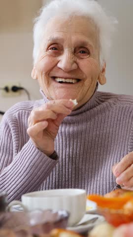Vertical video of happy smiling senior woman with bright open smile and sparkling eyes eating at table and drinking tea. Grandmother laughing surrounded by care and love. elderly woman happy