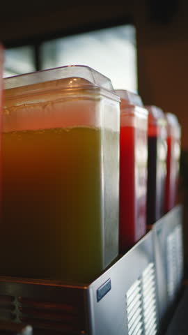 A slow, cinematic pan shot across a row of colorful fruit juices in beverage dispensers. Close-up with shallow depth of field highlights refreshing drinks in a cafe or buffet. Vertical video