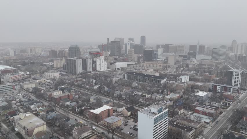 Drone moving backward while facing downtown Winnipeg Manitoba, showing buildings, roads, falling snow, and cloudy skies during early winter in the city.
