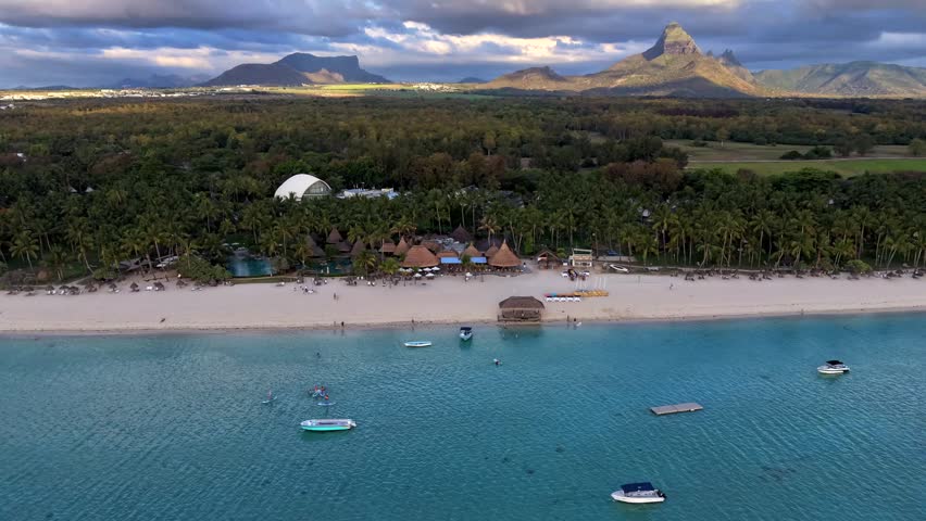 Aerial view shows turquoise sea. White sand stretches to shore. Boats float gently near beach. Lush green hills cover inland. Distant mountains under cloudy sky. People relax happily