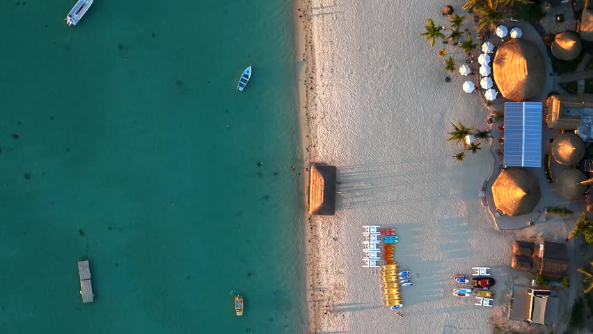 Aerial view of turquoise sea and white sand beach. Small boats drift gently on calm water. Bamboo huts dot the shoreline. People relax under umbrellas. Calm smiles fill the scene