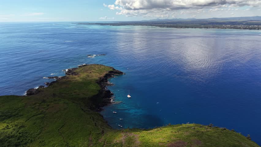 Aerial view of a lush green coastline. Deep blue ocean meets rocky shore. Partly cloudy sky above the scene. Calm waters highlight natural beauty. Perfect for nature tourism content