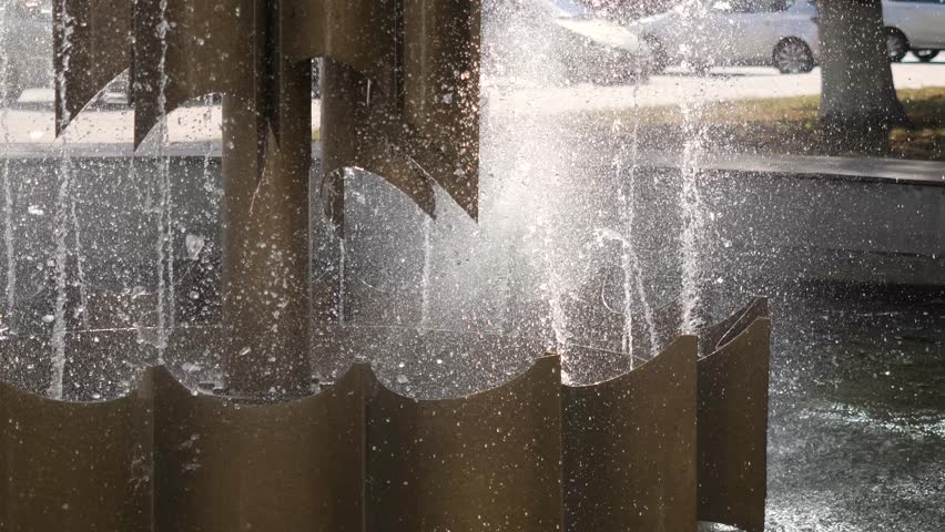 bronze colored multi-tiered metal fountain structure spraying glistening water droplets into air on sunny day in urban park
