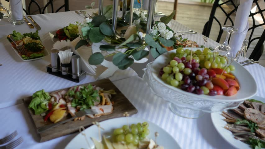 Elegant restaurant banquet table setup featuring variety gourmet appetizers including cheese platters with grapes honey meat delicacies fruit bowls floral centerpieces white tablecloth festive celebration setting
