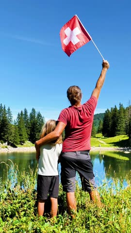 Rear view of father and son holding Swiss flag by mountain lake during summer vacation - 4k handheld shot