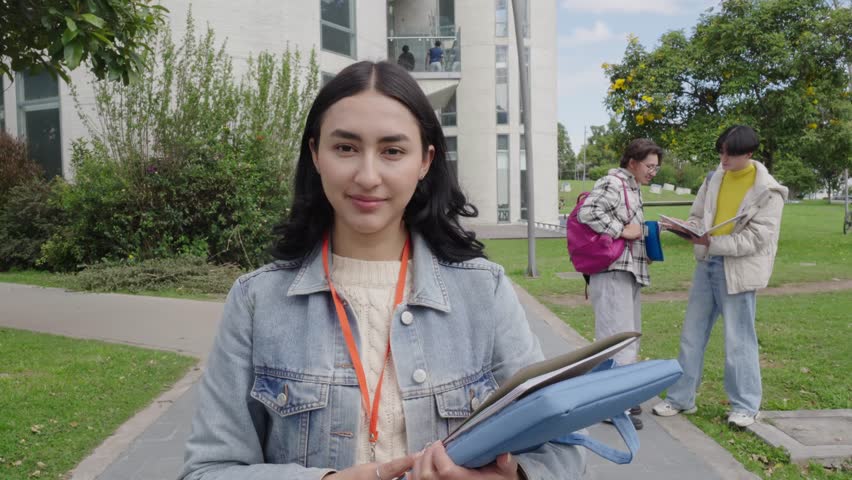Cheerful young woman holding a laptop case walking on the university campus path with other students talking in the background, she is looking at the camera and smiling on a bright sunny day