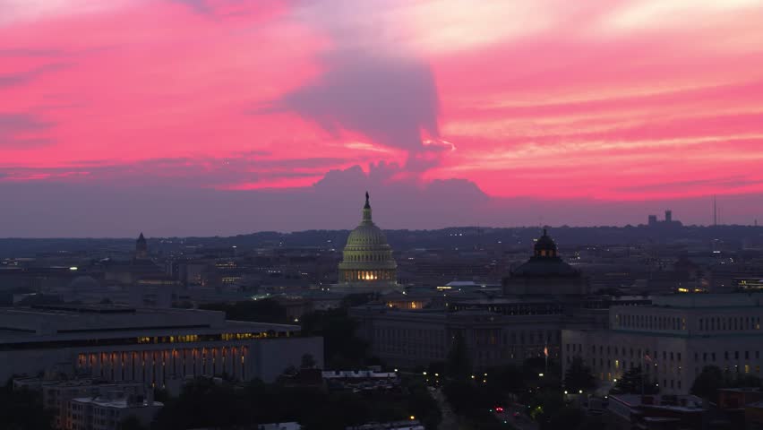Sunset drone shot of the U.S. Capitol with its illuminated dome, surrounded by city buildings under a vibrant sky.