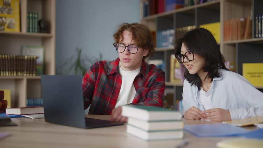 Two smart multiethnic college students working on a school project together using a laptop in a library, sharing ideas and collaborating to find information for their academic research