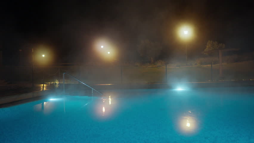 A serene outdoor swimming pool at night with warm steam rising from the inviting blue water. Illuminated by ambient streetlights