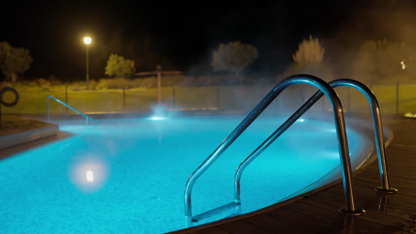 Static shot of an outdoor swimming pool at night, illuminated by blue lights, with steam rising from the warm water. Creates a serene, tranquil, and luxurious atmosphere for relaxation and spa