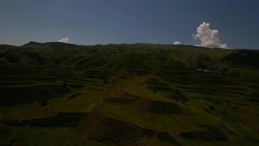 Aerial view of lush green hills in the Kavkaz region. The landscape features terraced fields and scattered trees under a clear blue sky.