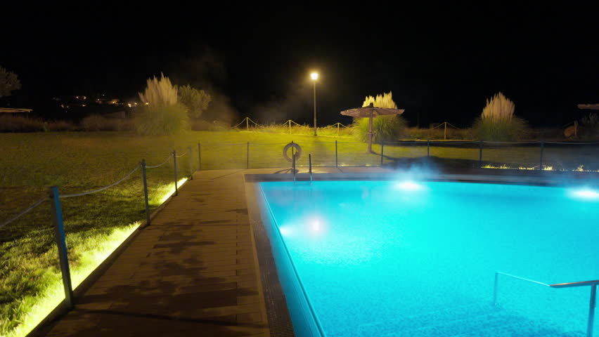 An outdoor heated swimming pool steams gently at night, illuminated by bright blue underwater lights and warm ambient lighting. The tranquil resort setting suggests relaxation and luxury