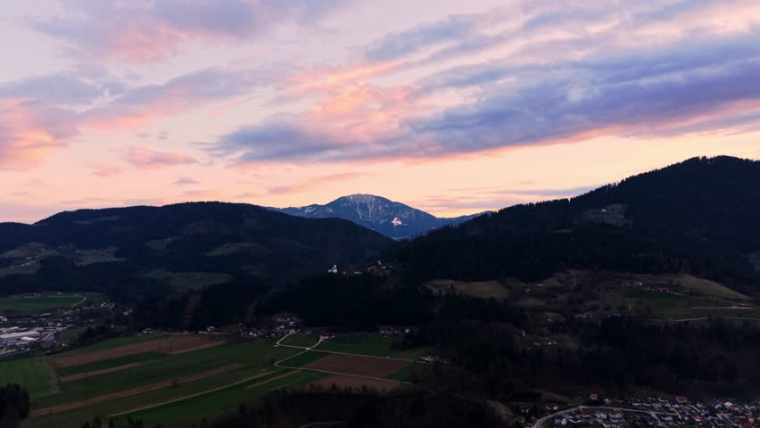 Aerial drone reveal of scenic mountain valley countryside with farmland, hills and colorful sunset sky in peaceful rural landscape.