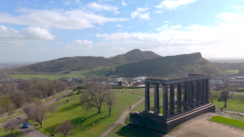 Smooth aerial orbit around iconic monuments on Calton Hill overlooking Edinburgh skyline at sunrise. Warm golden hour light illuminates historic architecture and cityscape, creating a cinematic travel scene ideal for tourism, documentary, and establishing shots.