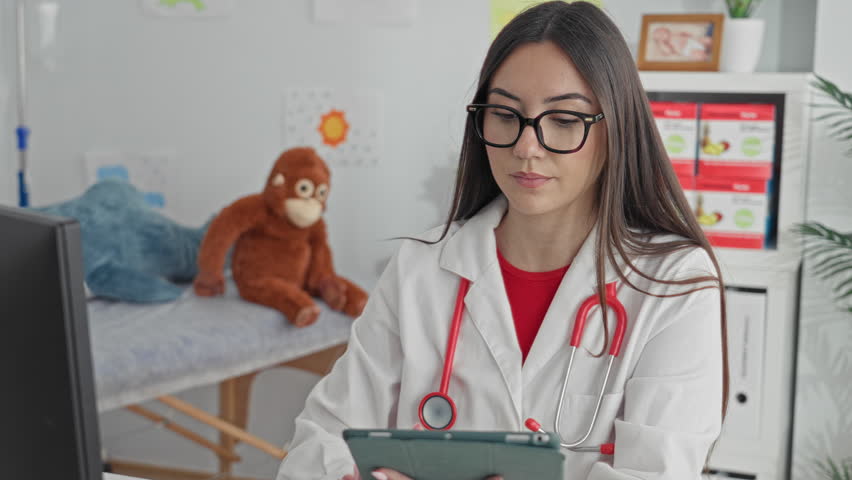 Woman doctor holding tablet and typing with hand, red stethoscope visible, pediatric exam bed and toys in clinic building; compassionate care.
