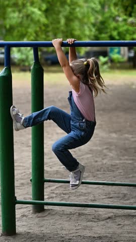 Little girl lifting legs while hanging on horizontal bar at outdoor playground. Girl performs active exercise in park, showing strength and energetic childhood lifestyle in summer setting. vertical