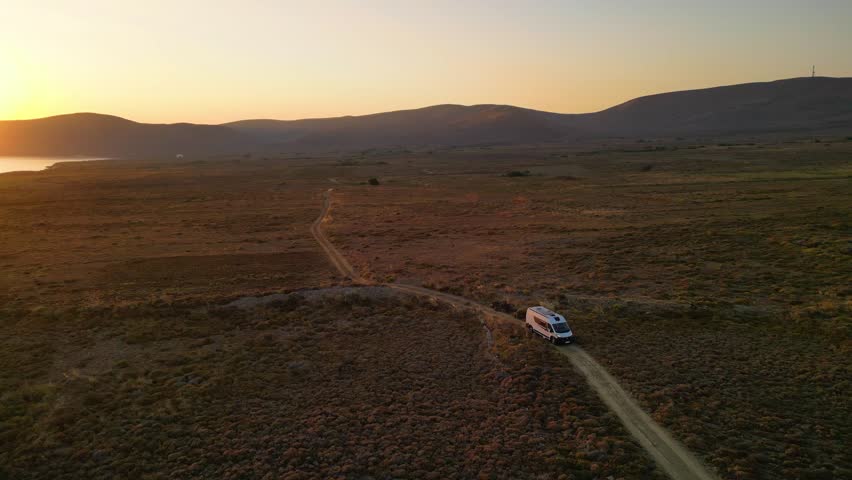 A camper van driving along a remote coastal road at sunset on Limnos Island, Greece. Aerial perspective shows golden Mediterranean light, rugged landscape and freedom of van life travel.