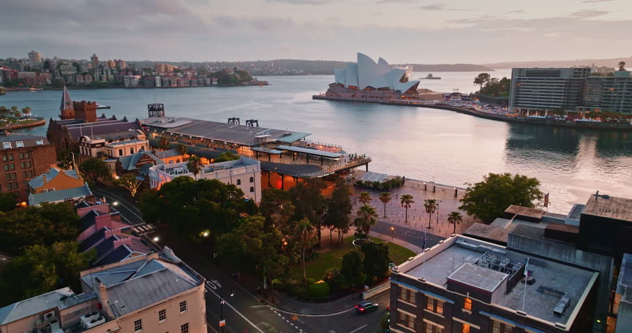 Sydney Opera House and Circular Quay waterfront at dramatic cloudy sunset dusk, ferries crossing the harbor and city skyline reflected in rippling water, iconic urban landmark. Aerial drone flight