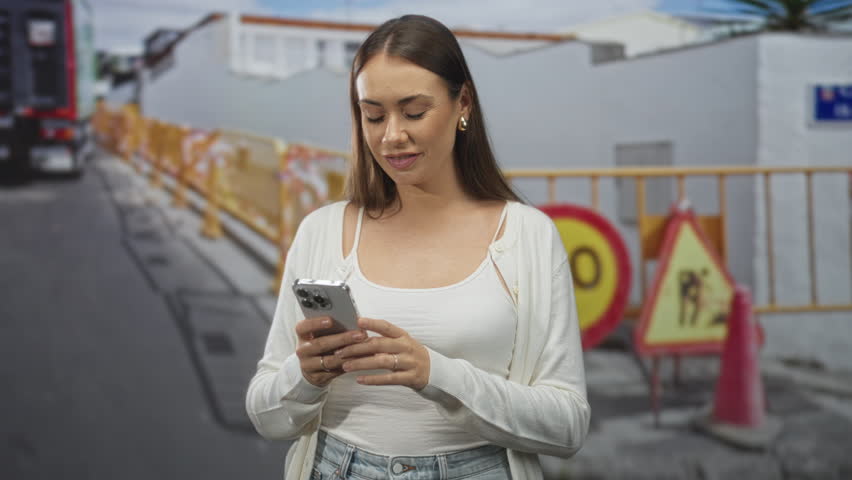 Woman holding smartphone, tapping screen and pointing index finger beside roadwork barricade on a street; focus everyday life.