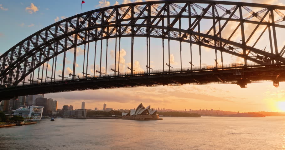 Drone flight under Sydney Harbour Bridge at sunset. Sydney Opera House, cruise ships, urban skyline reflecting warm colors across Sydney Harbour, vibrant atmosphere of bustling cityscape at twilight