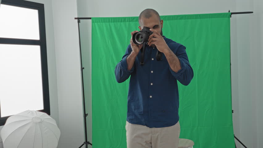 Young hispanic man smiling while holding camera, hands adjusting lens in studio; creative joy and inspiration.