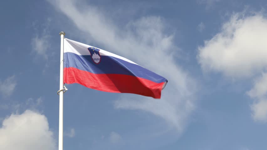 Slovenian national flag waving on flagpole against blue sky with clouds