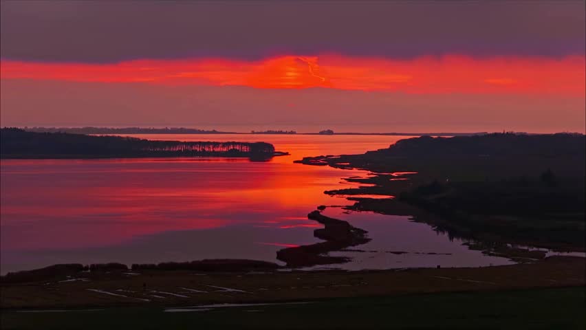 A vibrant and dramatic orange sunset illuminates the calm waters of a vast coastal landscape view