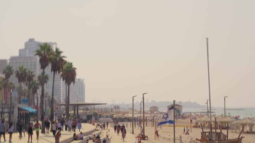 Tel Aviv promenade and beach as locals and tourists enjoy hot sunny day by sea. People swim walk and run by the Israeli coast on summer vacation holiday or weekend. Israeli flag waving in foreground