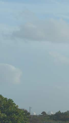A rocket launches vertically from a coastal space center with trees and infrastructure visible below. Bright engine flames rise into a cloudy blue sky as the spacecraft lifts off from the launchpad.