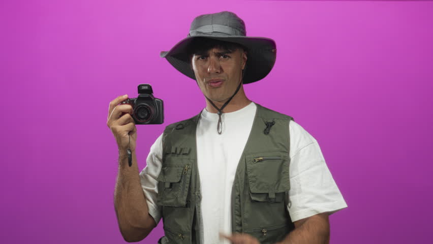 Man in explorer hat and vest holding camera and pointing finger at lens in studio; adventure confidence.